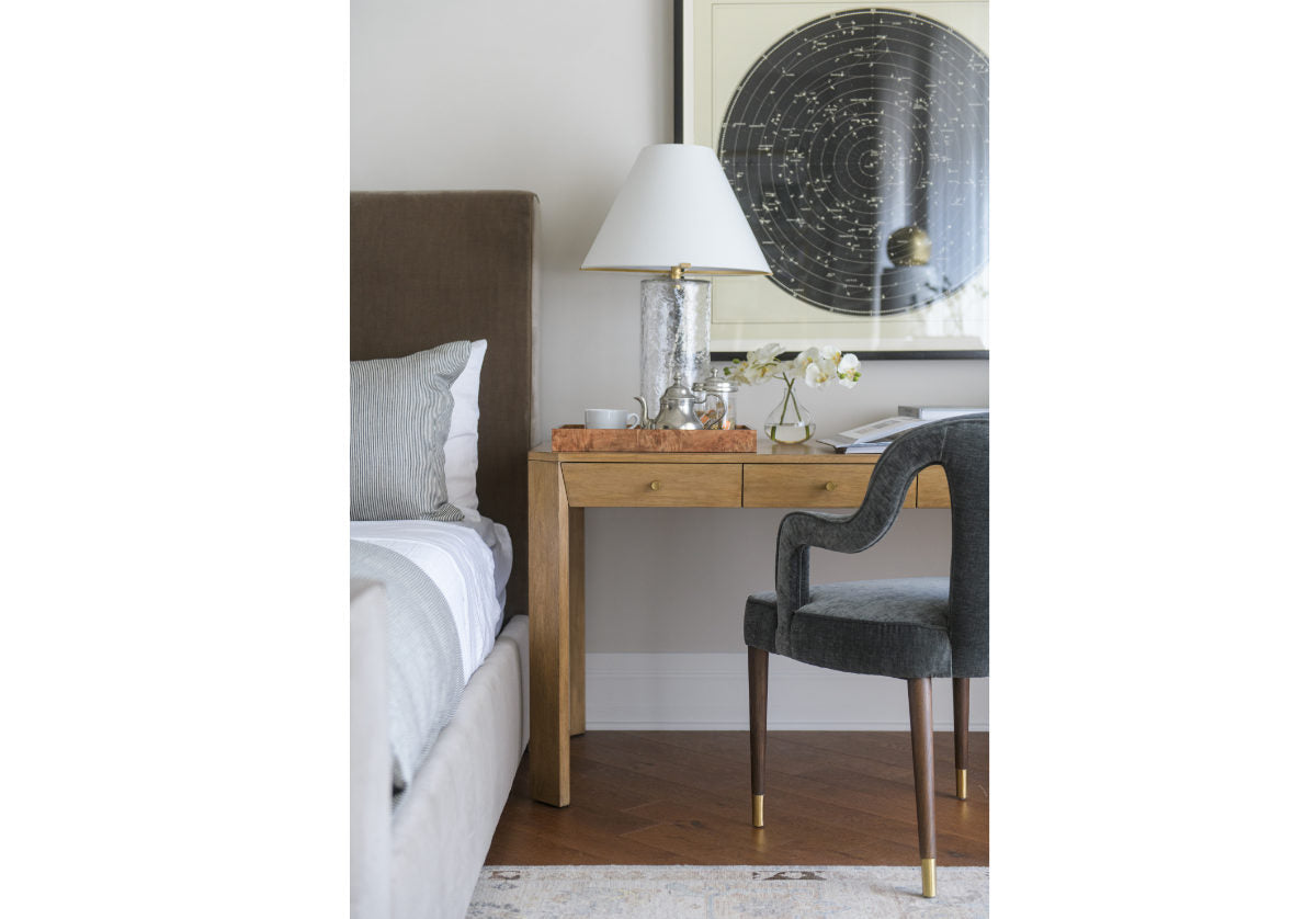 The Brooklyn Desk showcases a beautiful bleached walnut finish set on a walnut composite base. Its clean-lined from is subtly contrasted by angled drawers with petite bronze knobs for added dimension and sophistication. Easily style at the foot of a bed or in an office for an eye-catching design. with the beautifully designed bobbie chair placed in front. decorated with a silver tea pot and burled wood tray next to a bed.