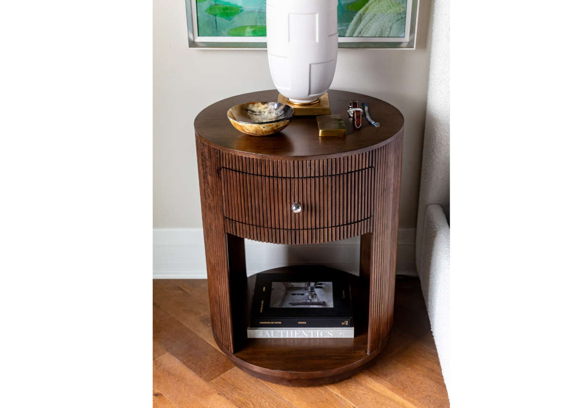 THE AUTHENTICS by Random House Inc is a round wooden side table with vertical grooves, featuring a drawer with a knob. A decorative bowl, lighter, keys, and lamp adorn the top while two stacked books rest below—a tastemakers favorite.