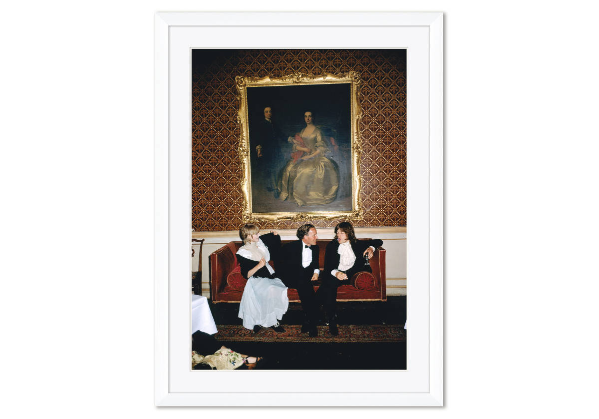 picture of mick jagger smoking and two other people enjoying his company while sitting on a couch with a large painting hanging above them.