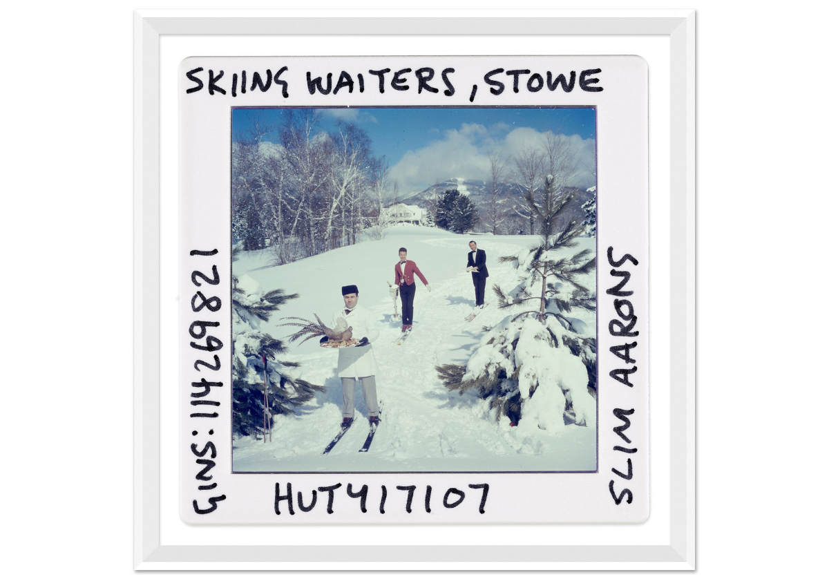 three men dressed as waiters as they ski down a hill hooding items. writing on the sides of the picture can be seen.