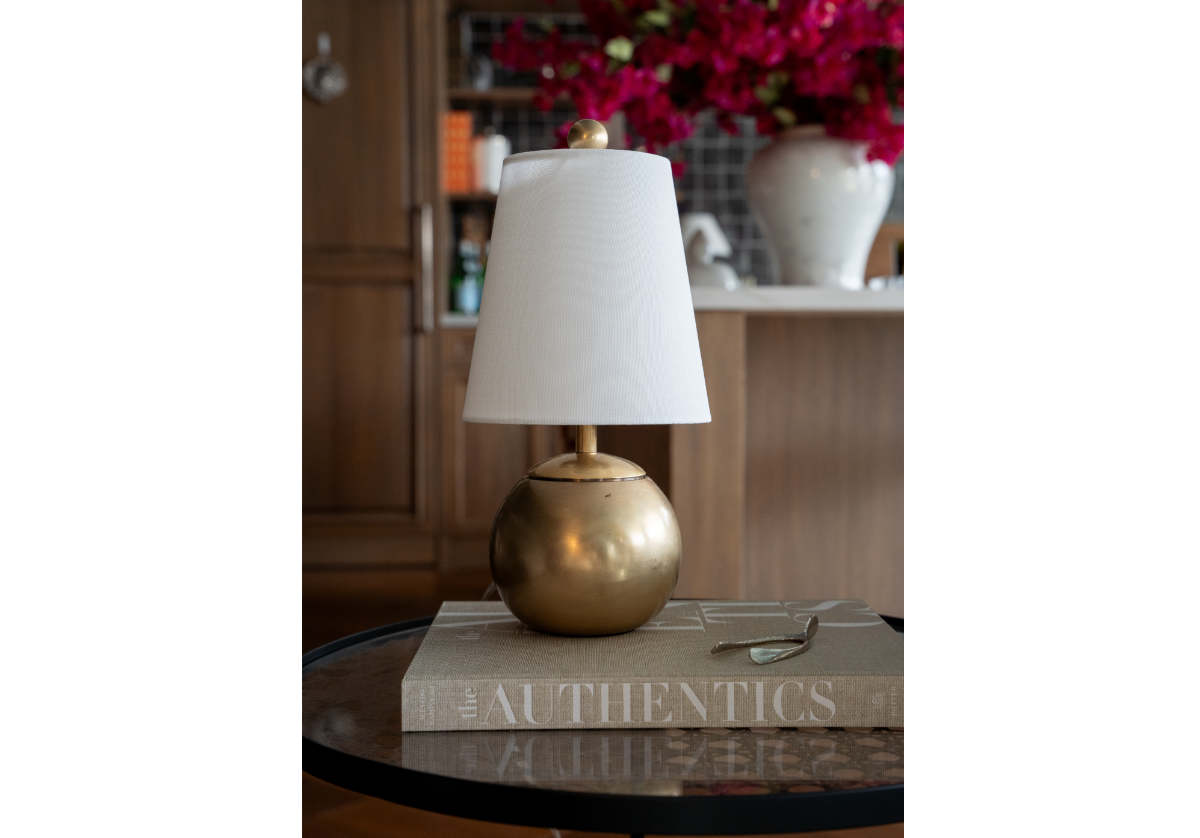A small lamp with a white shade and round gold base sits on THE AUTHENTICS by Random House Inc, nodding to tastemakers. This setup graces a round table accompanied by a spoon, with blurred cabinets and vibrant flowers in the background completing the scene.