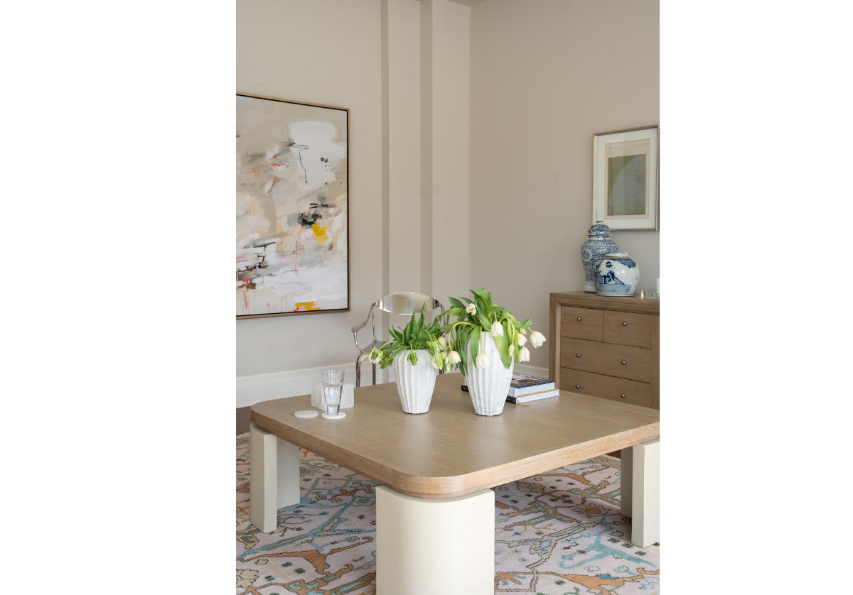 lifestyle photo of the blair cocktail that makes a sculptural statement with its spacious white oak top elevated by four radius legs. The natural wood graining and patterning provide a beautiful contrast to its smooth, creamy base for an elevated design. there is a beautiful rug in the center of the photo with a dresser and jars atop of it.