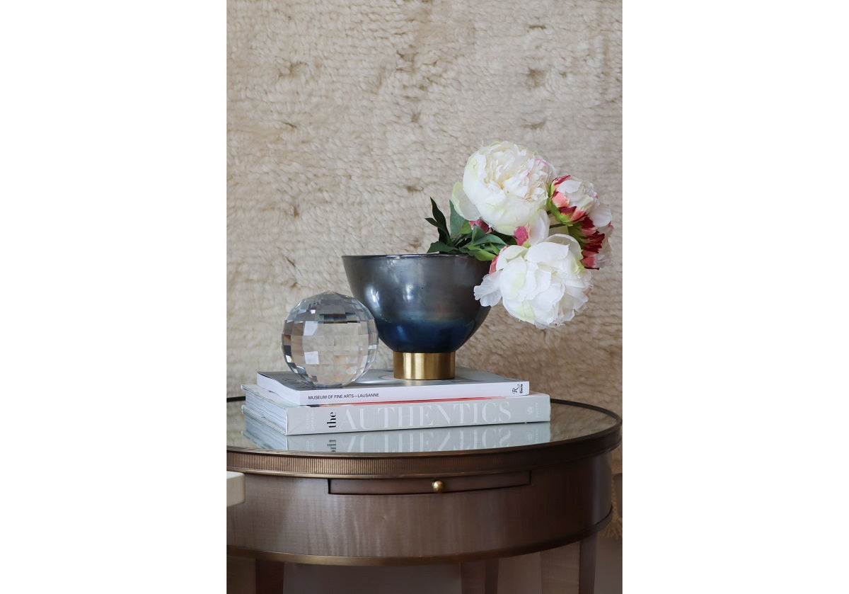 THE AUTHENTICS by Random House Inc sits on a small round table adorned with a metallic vase of white and pink flowers. A clear sphere complements the display, set against a textured fabric wall that exudes elegant style.