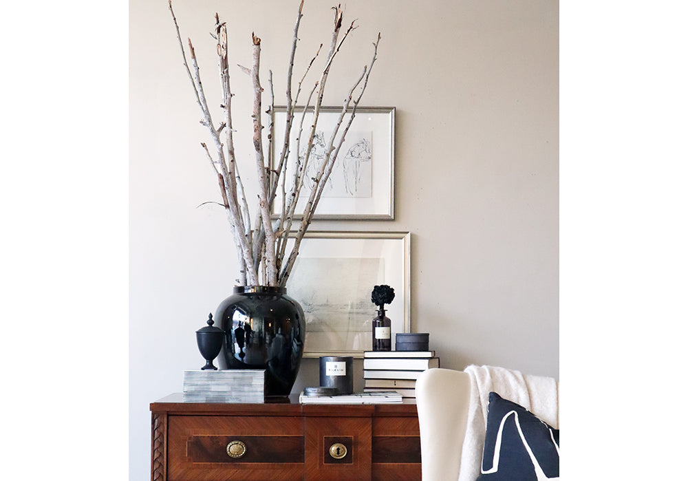 lifestyle photo of a large black vase on a dresser with books. the black vase has sticks coming out of it, giving a lovely feel to the room.