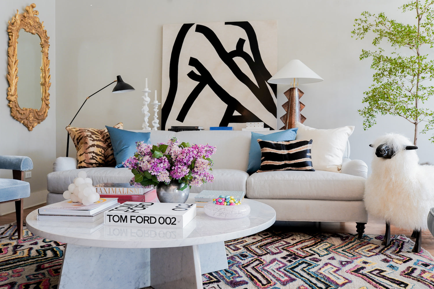 A grey couch is in the middle of the living room. There are two floor lamps on either side of the couch. On the left is a blue velvet armchair. On the right is a light brown leather armchair with a white circular ottoman.