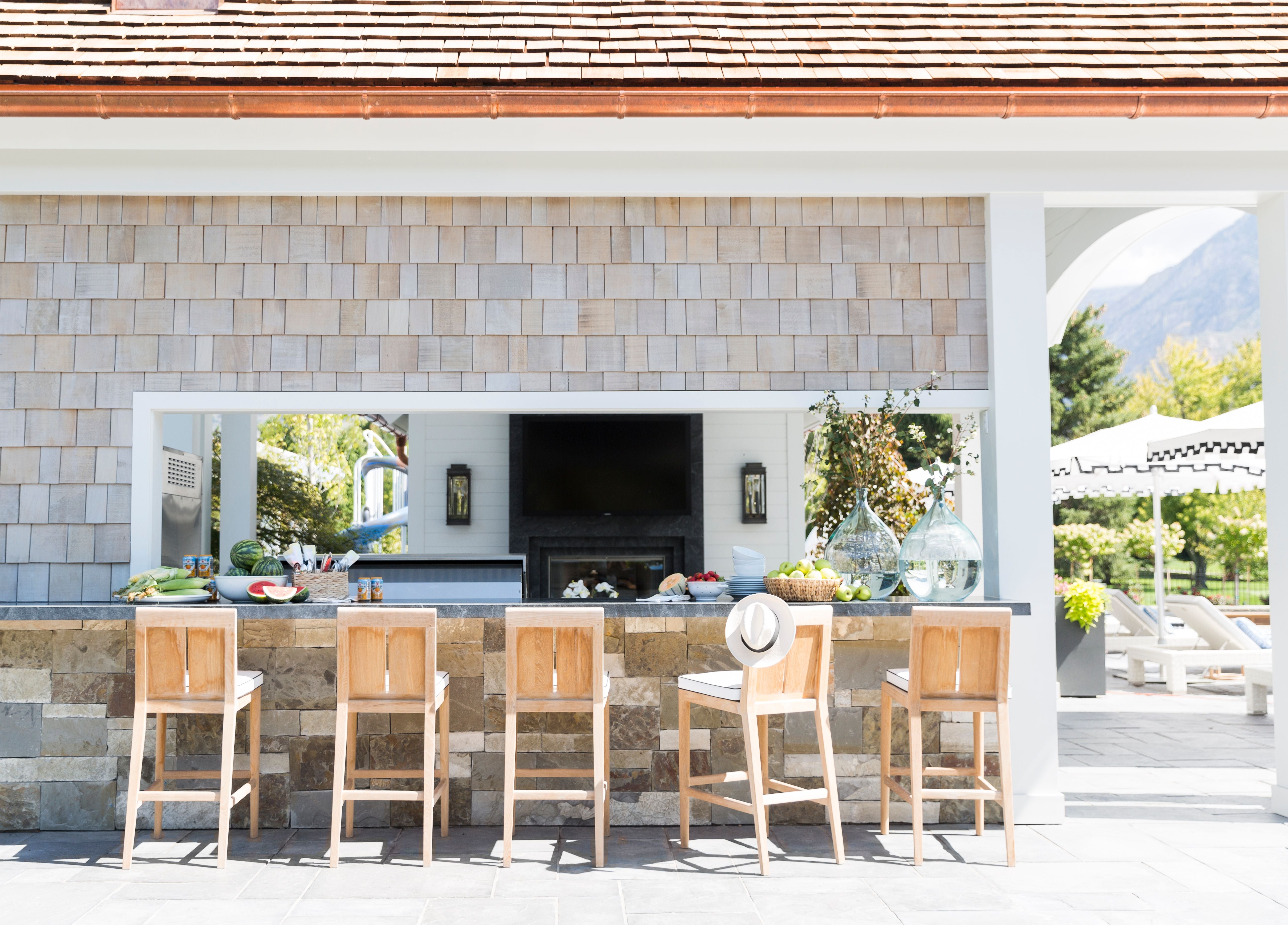 There is a pool house with five wooden bar chairs at the counter. To the right are umbrellas with pool lounging chairs. Inside the pool house are lanterns and a fire place.