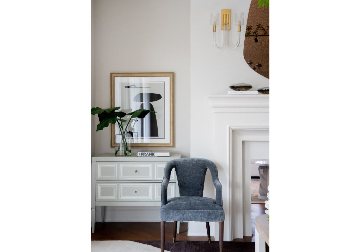 a lifestyle photo of the bobbie chair, sitting in front of a fireplace mantle in front of a white and gray dresser with a palm stem on top of it. 