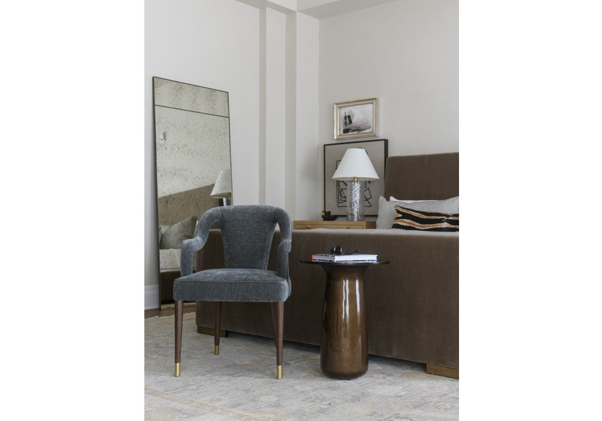 The Bobbie showcases a shapely form with its sloping back, curved arms, and canted legs. Its subtle textured upholstery in a smoky aegean adds a sophisticated style. Brass-capped feet beautifully contrast its walnut-finished legs for a luxurious, tasteful design. the monroe bed can be seen in the background of the photo with a large eden floor mirror. 