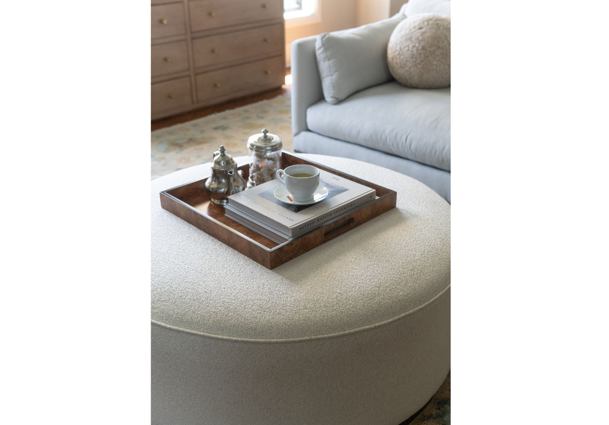 a lifestyle photo of the piper ottoman with the burled wood tray with books and a tea pot inside of it. 