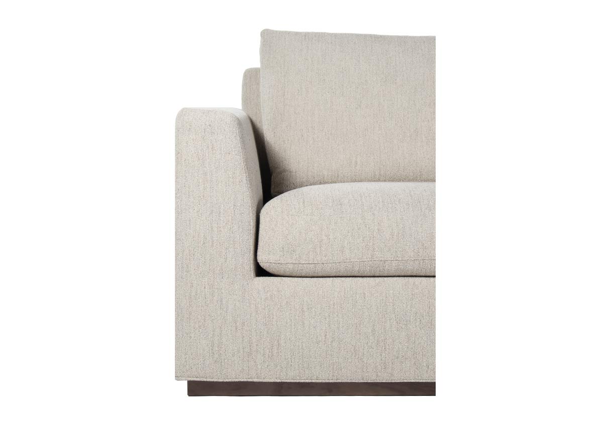 close up of august sofa corner. The angled arms and walnut-stained plinth base add dimension and interest making this piece beautifully suited for spacious living or family rooms. Its neutral woven fabric is subtly accented with a blue loop undertone for added depth