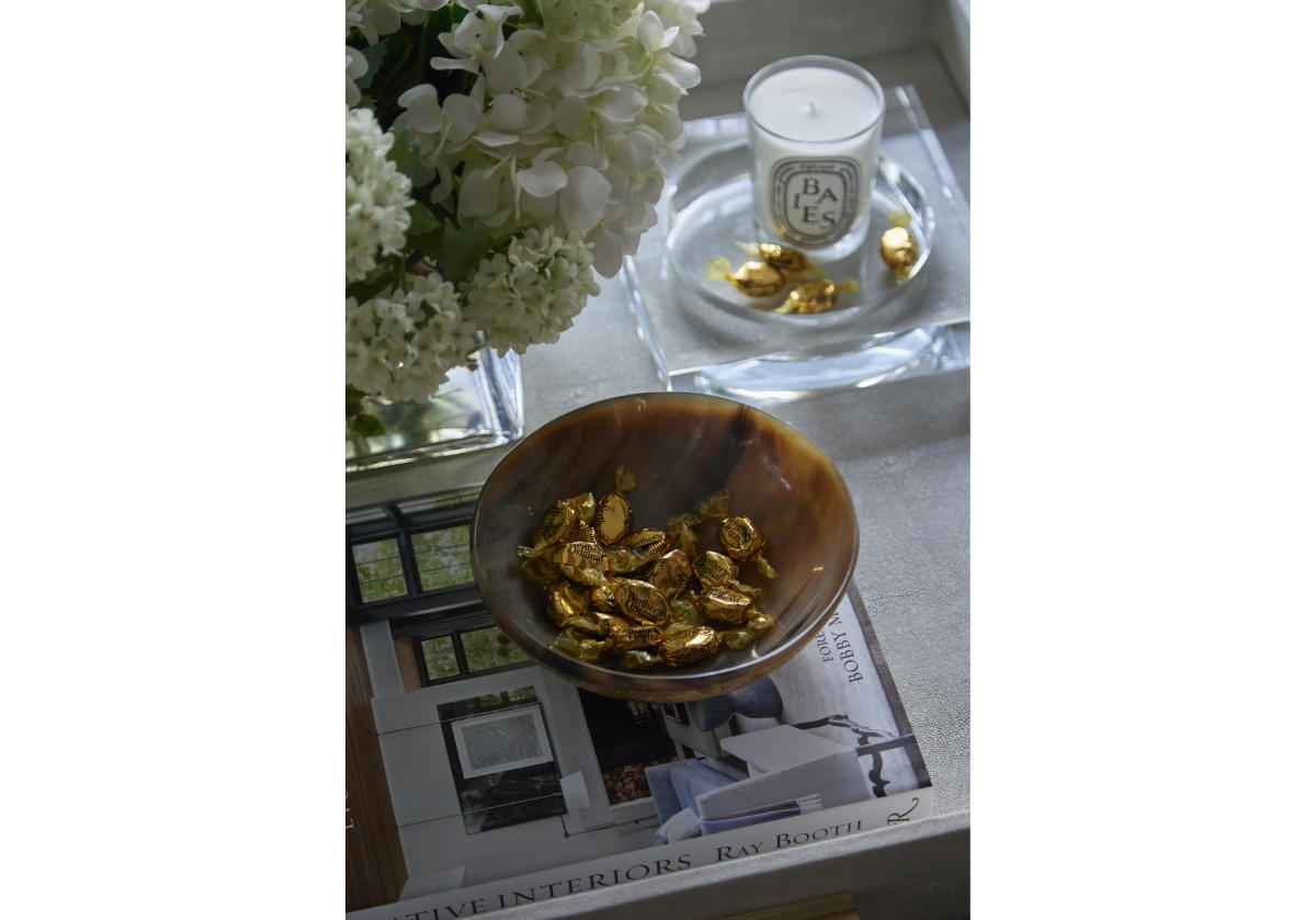 Next to a large bouquet of flowers on a coffee table, there is a crystal candle dish with caramels inside of it and a bone bowl filled with more candies next to it on some books. 