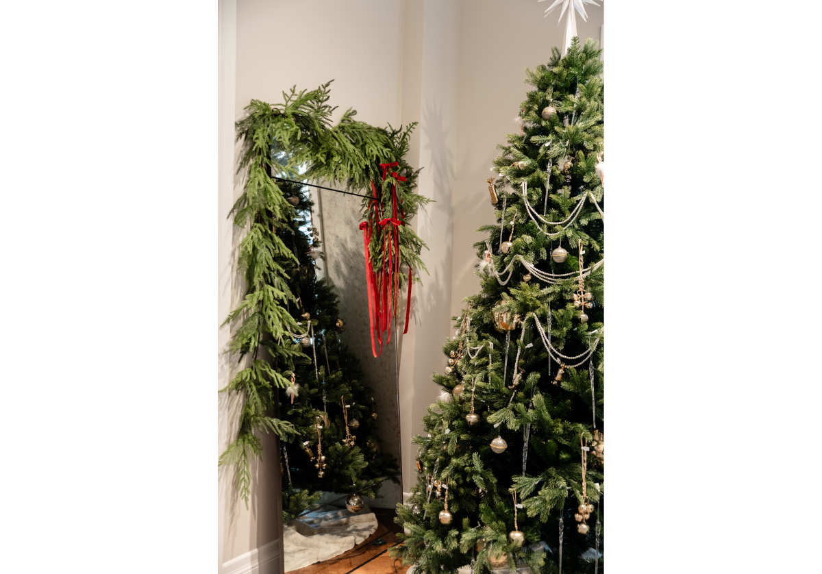 eden floor mirror with a pine garland and red ribbon draped over it with a christmas  tree in the reflection. 