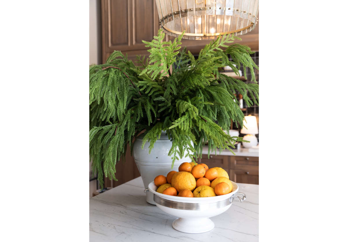 fir branches inside of a white vase with a bowl of oranges next to it. 