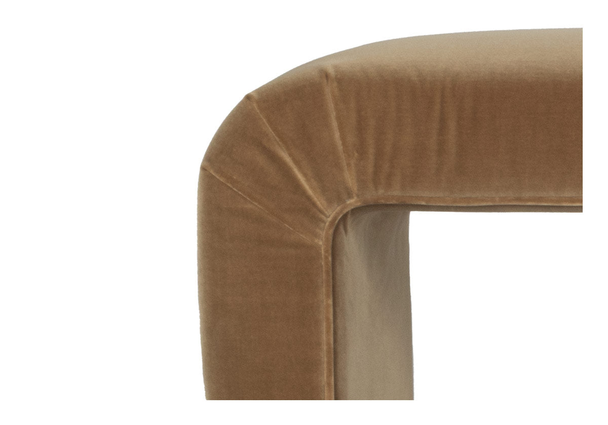 a picture of the corner of the phoebe bench is a luxurious italian cotton velvet chair, adding a touch of elegance and sophistication to any home. A soft brown suede colored velvet bench. The clean lines and sculptural form create a striking visual impact, while the curved edges offer a comfortable and inviting seat. Perfect for adding a pop of color and a touch of vintage flair to your living room, bedroom, or entryway.