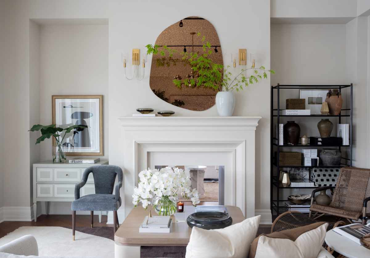 a lifestyle photo of the phoenix mirror hanging above a fireplace mantle. to the left there is a gray and white dresser. to the right there is a black shelf. in the center of the photo is the blair cocktail table. the the left of that is the bobbie chair. 