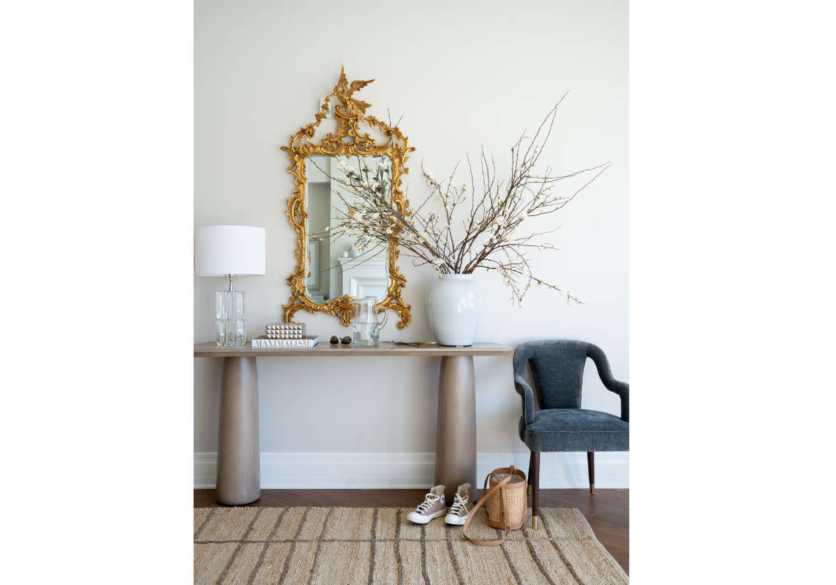 a lifestyle photo of the bobbie chair with the cate console with a large white vase and stems on top. there is a large decorative mirror above the cate console and the left of that is a crystal lamp. 