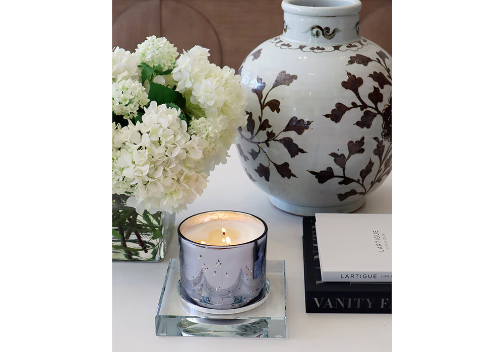 lifestyle photo with a candle inside of it and a large vase with flowers behind it. 