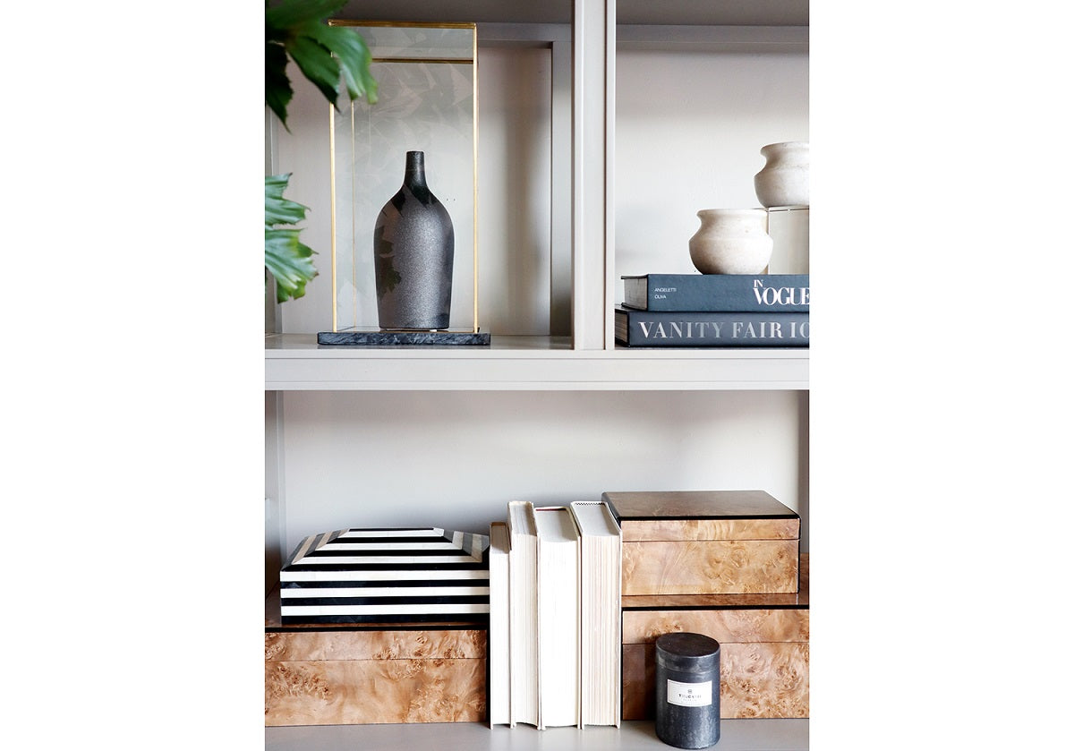 a lifestyle photo of the brass glass display box with a bottle inside of it on some shelves. pictured with other alice lane products like the burled box. 