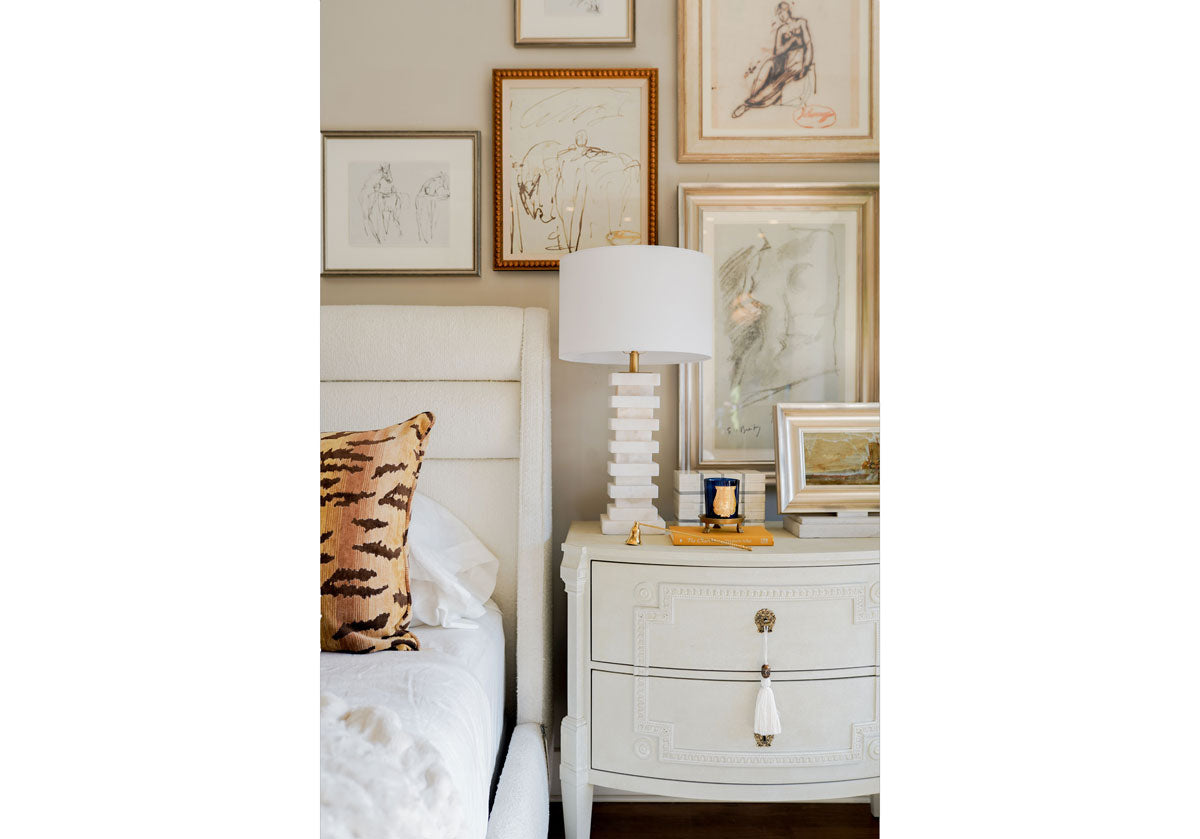 lifestyle photo of the chester lamp sitting on a dresser with beautiful sketches framed behind it. sitting next to a white bed with a tiger pillow. 