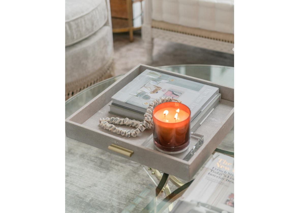 There are some books, a lit candle, and bone beads placed inside a decorative tray on a glass table. 