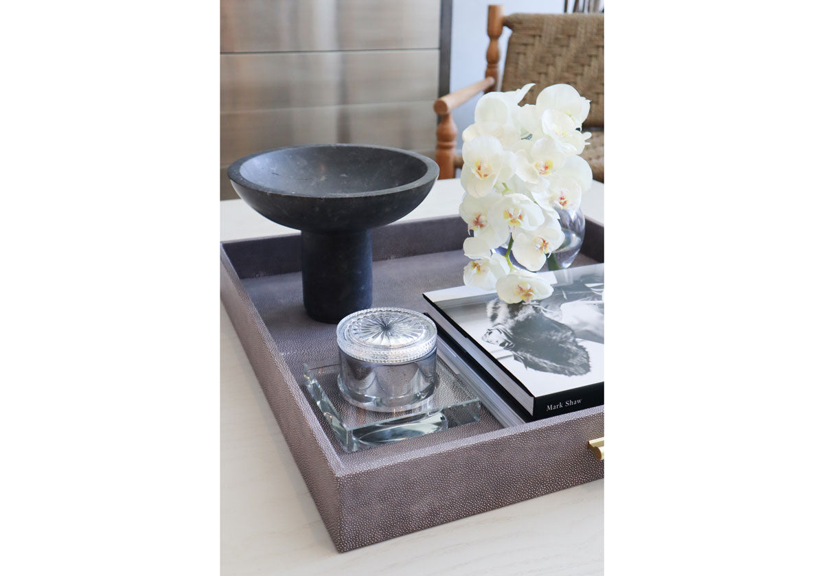 Inside of a gray tray is a crystal candle dish with a candle inside of it and books with flowers. 