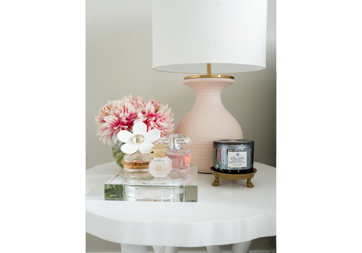 a close up of the crystal candle dish with perfumes inside of it and a light pink lamp in the background on a white side table. 