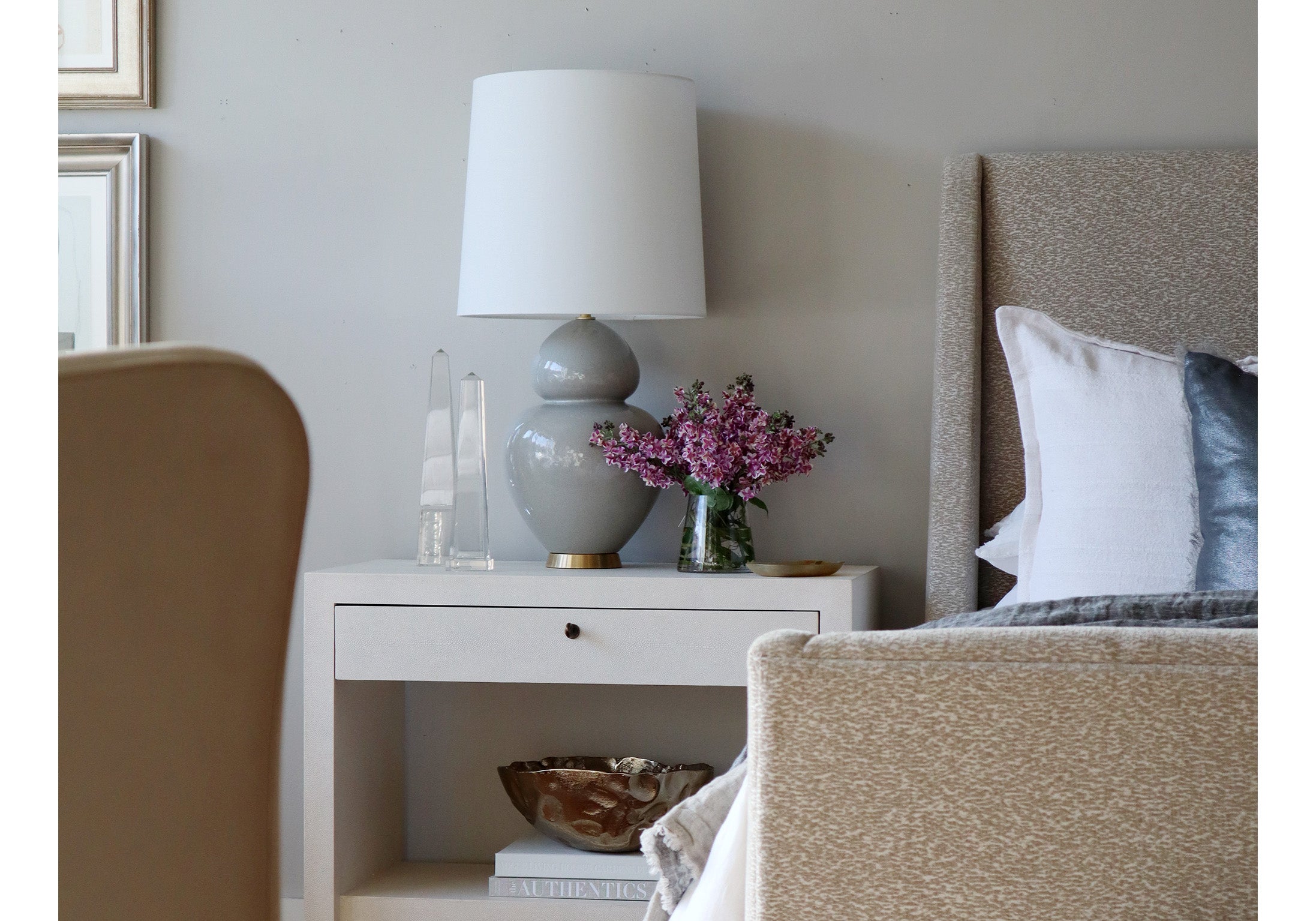 two different sized obelisks are seen on a table next to a vase of flowers and a gray lamp on a white side table next to a bed. 