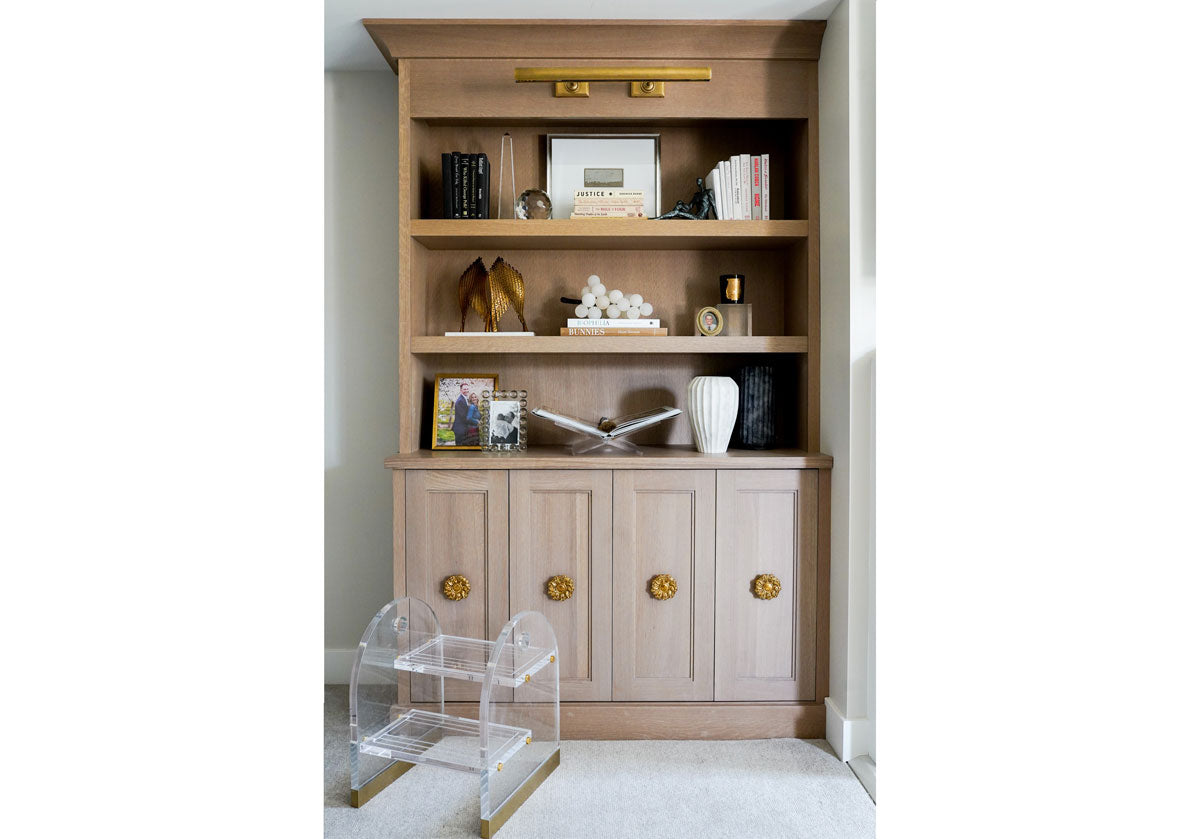 on this large wood shelf there is a crystal obelisk to the left on the the top next to a crystal orb, framed picture and books. underneath that shelf is a gold eros statue, marble grapes. underneath that shelf is alice lane's crystal orb frame with a lucite book display. on the floor is the lucite odette stool. 